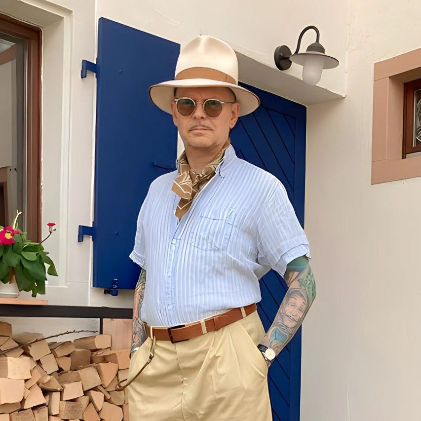 Fashion-forward man in a chic cream fedora, accessorized with a bandana and sunglasses, posing confidently outside a house with a vibrant blue door.