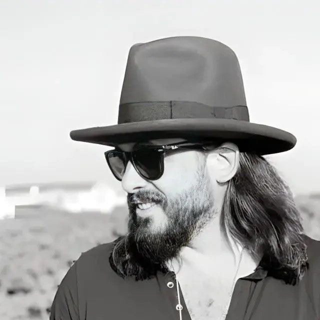 A stylish man in a custom black hat and sunglasses looks away thoughtfully, embodying modern elegance and personal flair against a serene background.