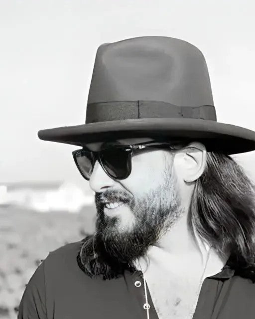 A stylish man in a custom black hat and sunglasses looks away thoughtfully, embodying modern elegance and personal flair against a serene background.