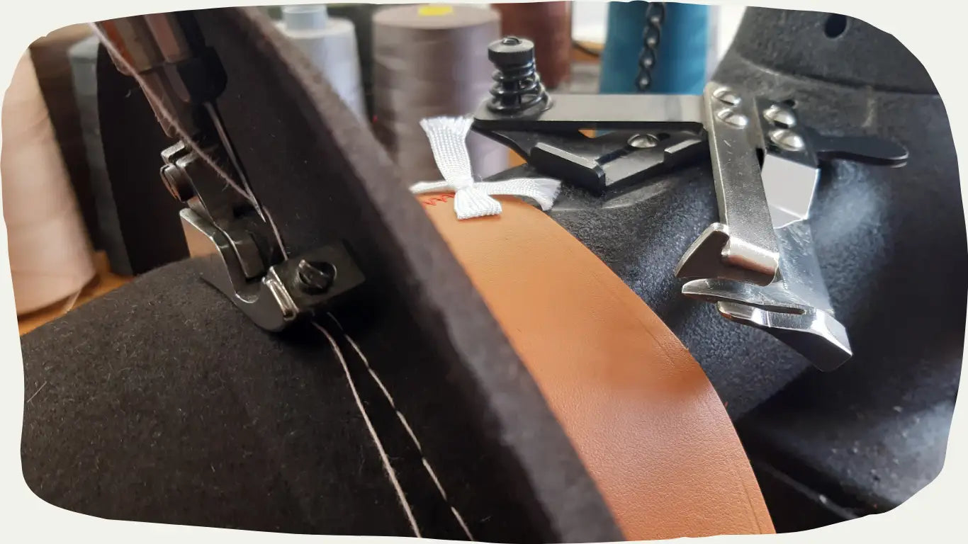 Close-up view of a hat being crafted with a sewing machine, highlighting the meticulous work of adding a leather band to the hat's base.