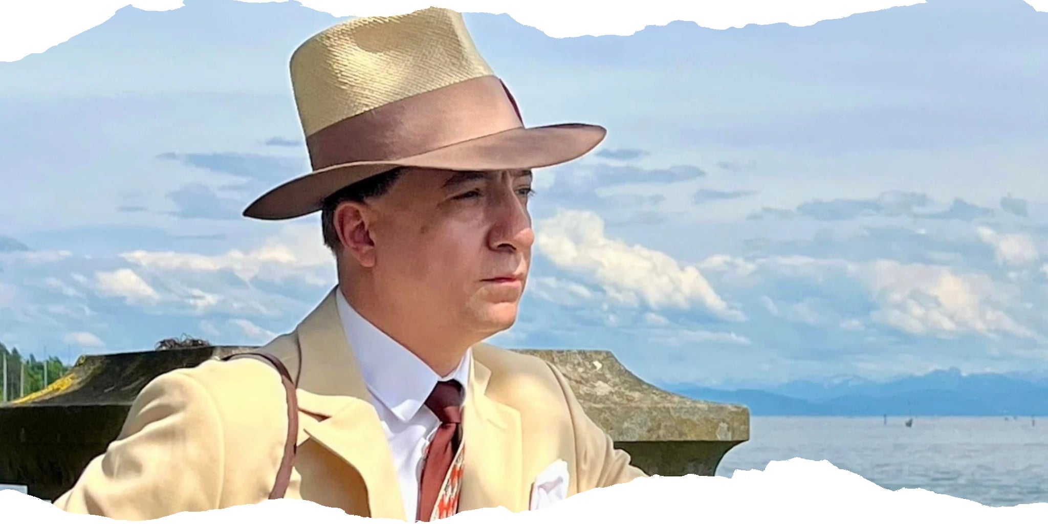 Elegant man in a cream suit and double-tone straw felt Panama hat, gazing thoughtfully by the lakeside with distant mountains in the background, exemplifying classic summer style.