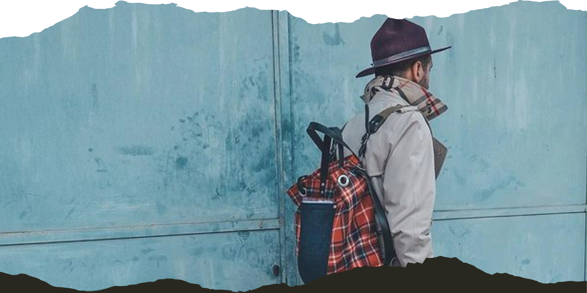 Man in stylish attire with a burgundy hat and backpack standing against a teal wall, facing away.