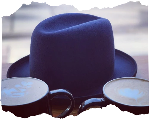 Stylish navy blue hat placed on a table next to two cups of coffee with artful foam, creating a relaxed and fashionable atmosphere.