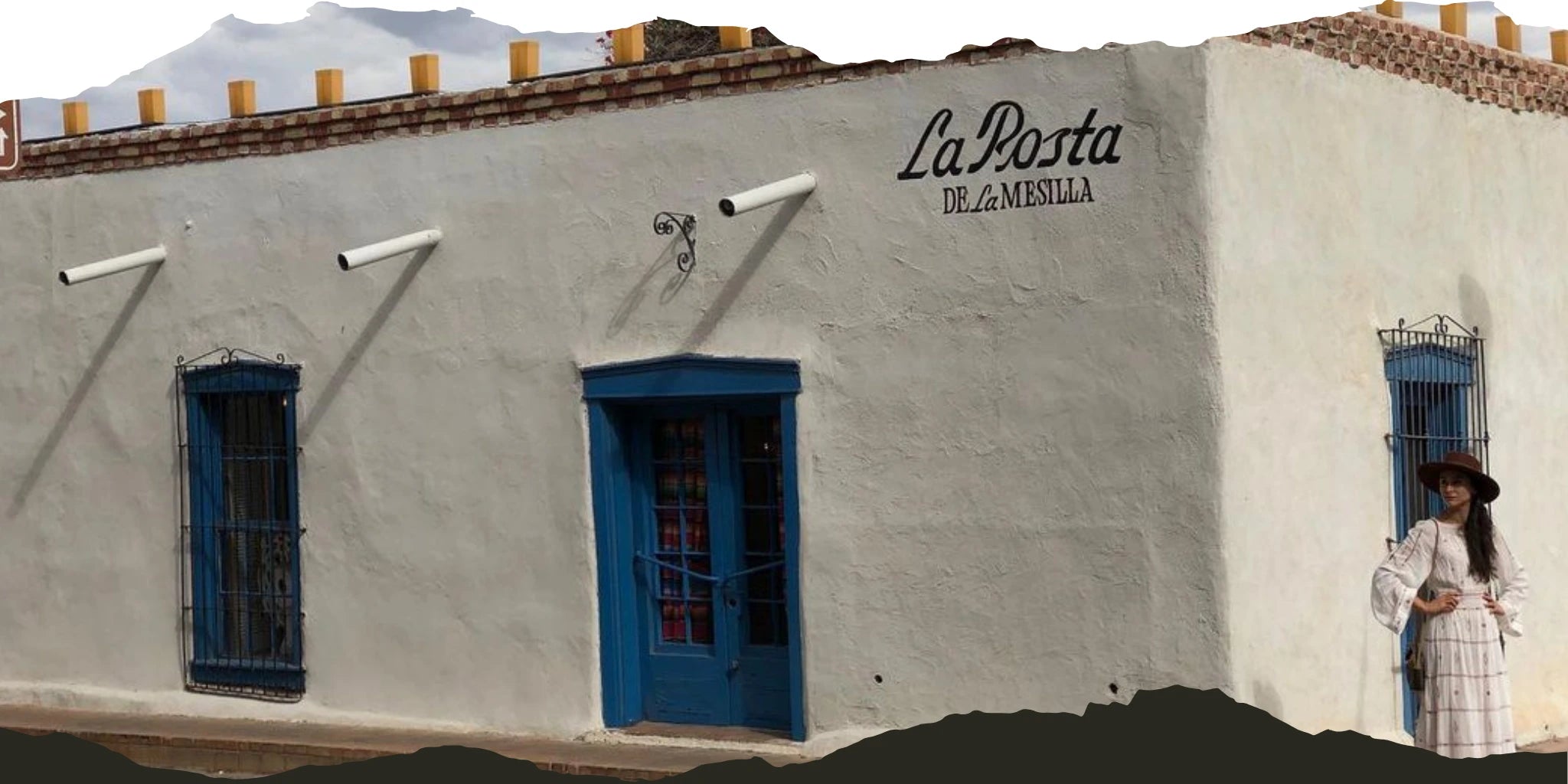 Exterior of La Posta de La Mesilla with a woman in a wide-brimmed hat and vintage-style dress standing near the blue door, evoking a rustic and timeless aesthetic.