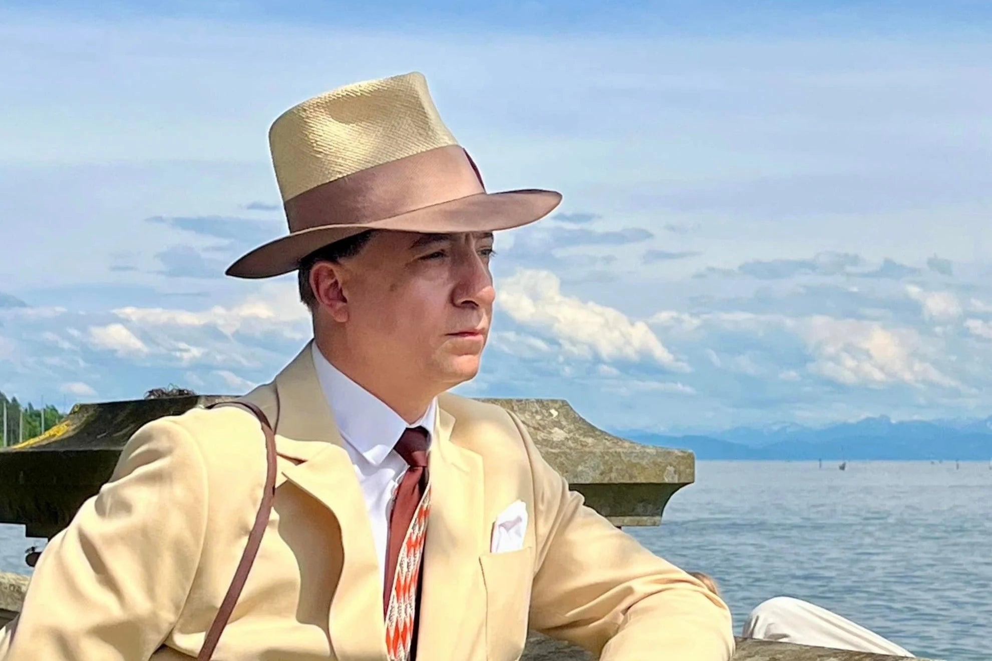 Elegant man dressed in a cream suit with a stylish Panama fedora hat, looking thoughtfully across a serene lake with distant mountains under a blue sky.