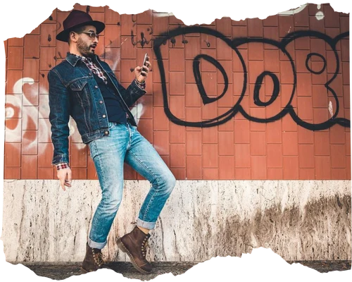 Stylish man in a cowboy hat and denim jacket leaning against a graffiti-covered wall while checking his smartphone.